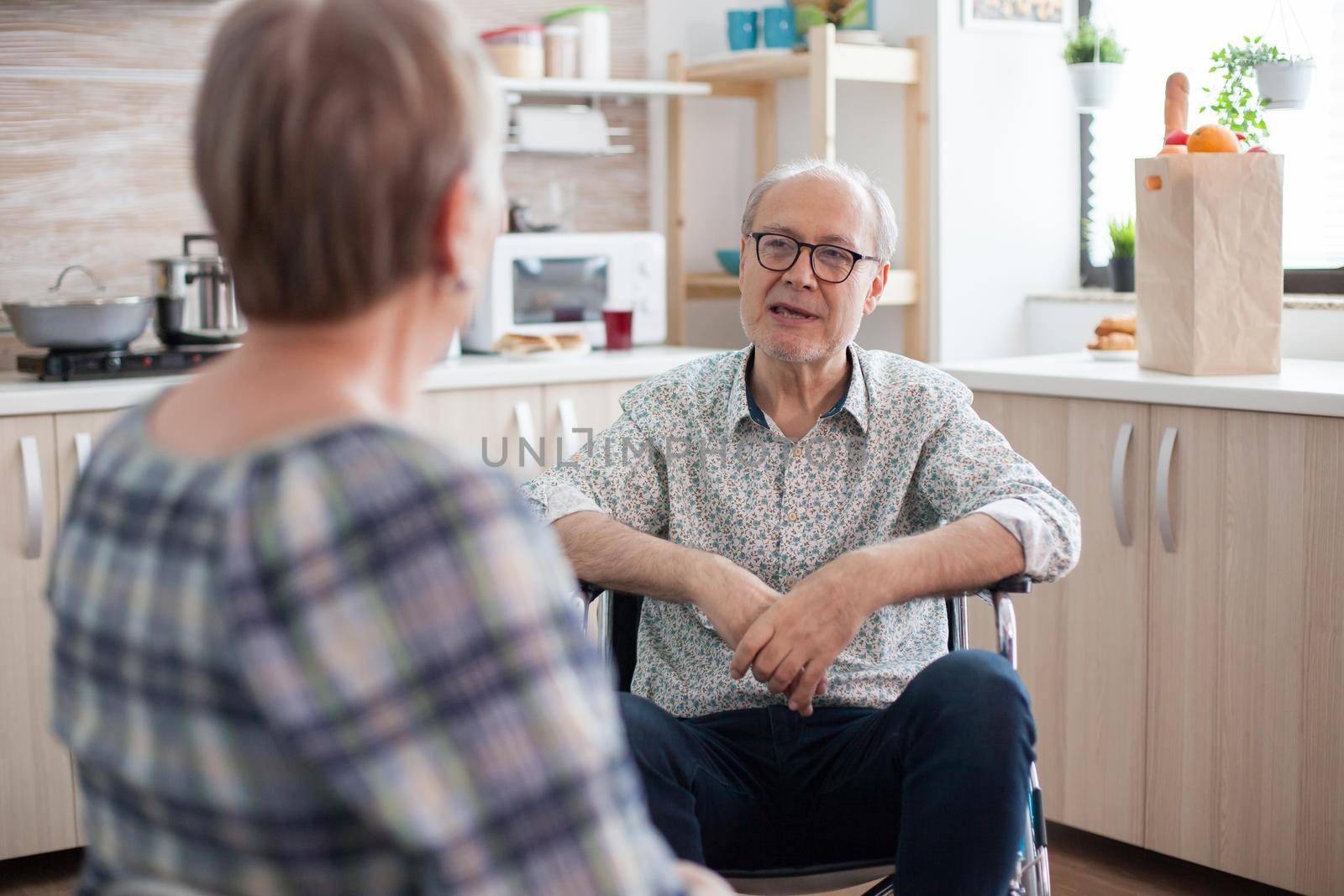 Disabled senior man talking with wife in the kitchen. Elderly person having a conversation with husband in kitchen. Living with disabled person with walking disabilities