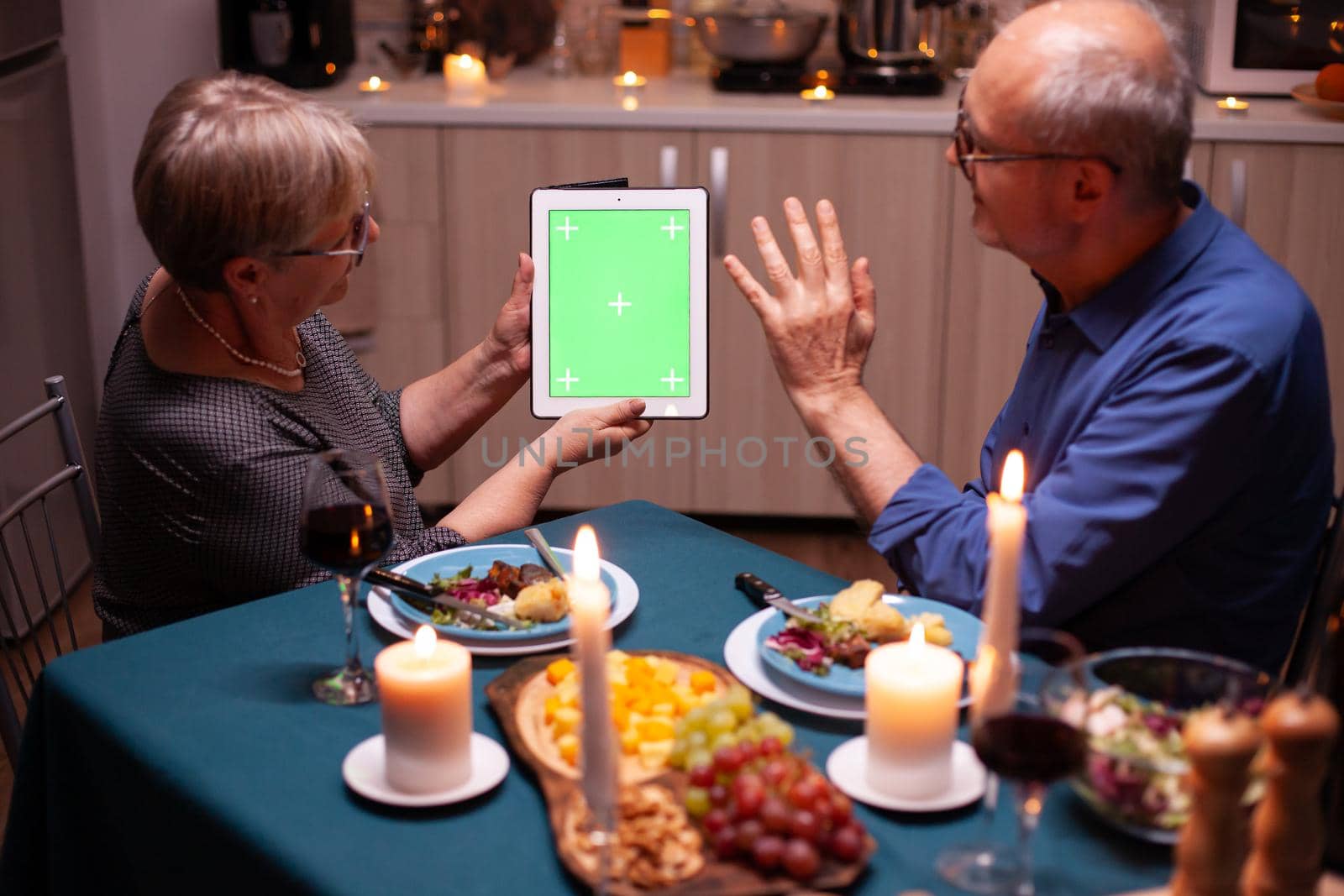 Elderly wife and husband waving at tablet pc with green screen. Aged people looking at green screen template chroma key display sitting at the table in kitchen during dinner.