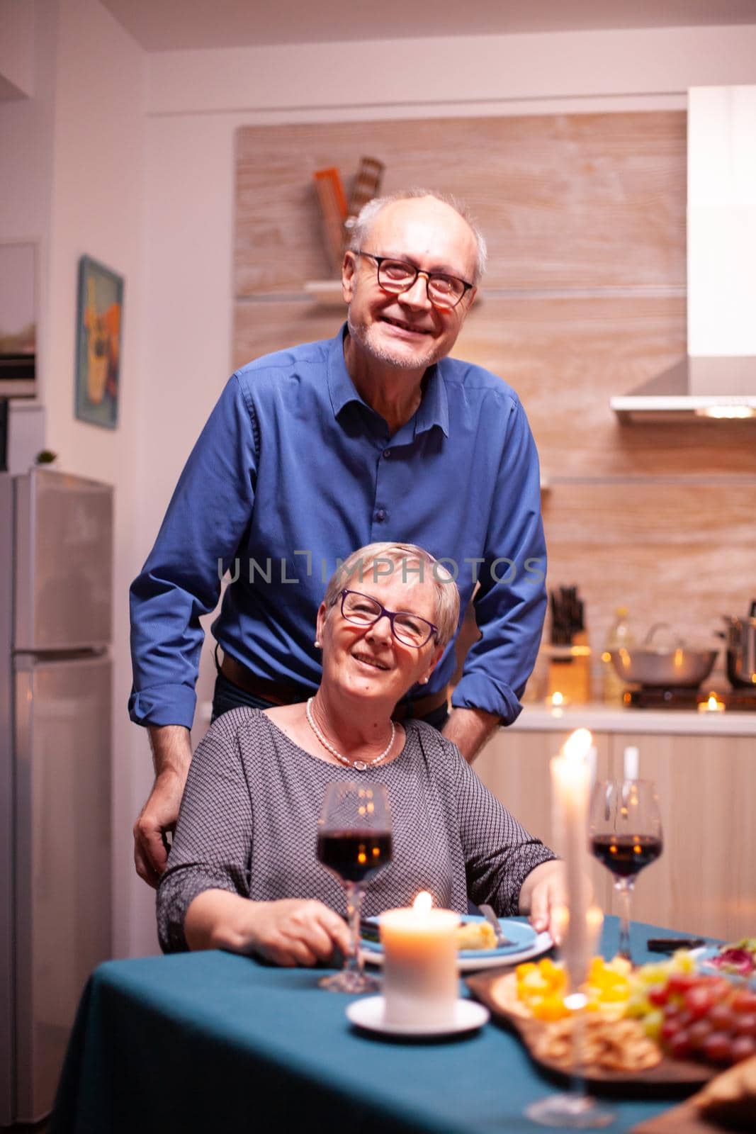 Disabled senior woman in wheelchair and husband looking at camera. Happy cheerful senior elderly couple dining together in the cozy kitchen, enjoying the meal, celebrating their anniversary.