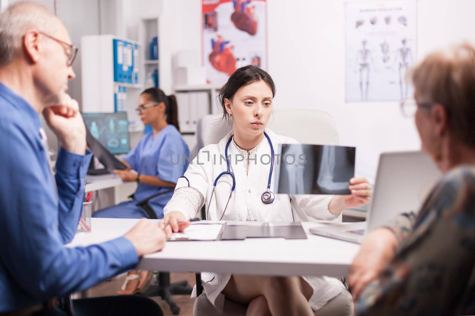 Doctor examining elderly man radiography during consultation in hospital office. Radiologist wearing white coat and stethoscope. Nurse in blue uniform.