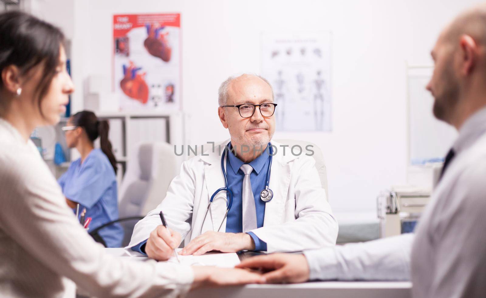 Worried couple holding hands during medical check in hospital office. Senior doctor with grey hair wearing white coat and stethoscope during examination.