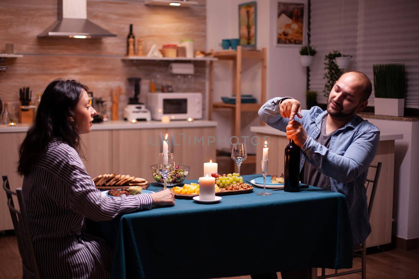 Cheerful husband opening a bottle of wine during romantic dinner with wife in kitchen. Happy couple talking, sitting at table in kitchen, enjoying the meal, celebrating their anniversary .
