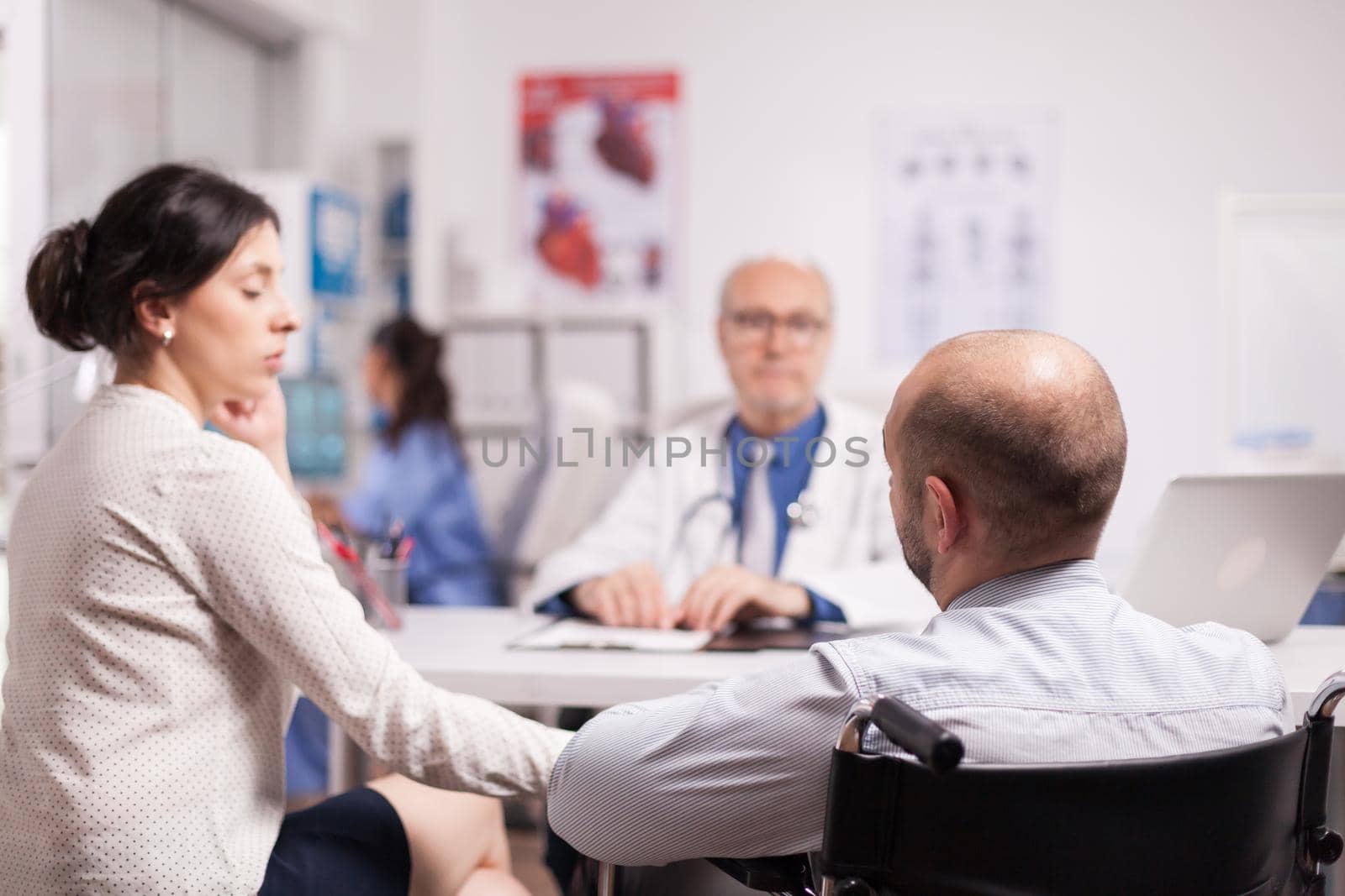 Disabled young man in wheelchair and his wife during consultation with senior doctor in hospital office. Elderly aged doctor wearing white coat and stethoscope. Nurse with blue uniform in the background.