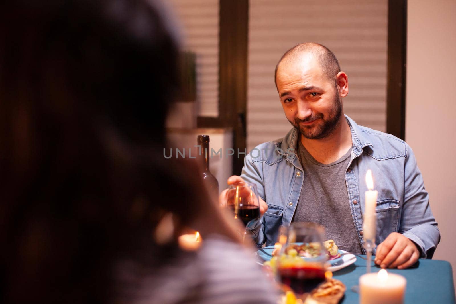 Man looking at wife while having romantic dinner and holding glass with red wine. Talking happy sitting at table dining room, enjoying the meal at home having romantic time at candle lights.