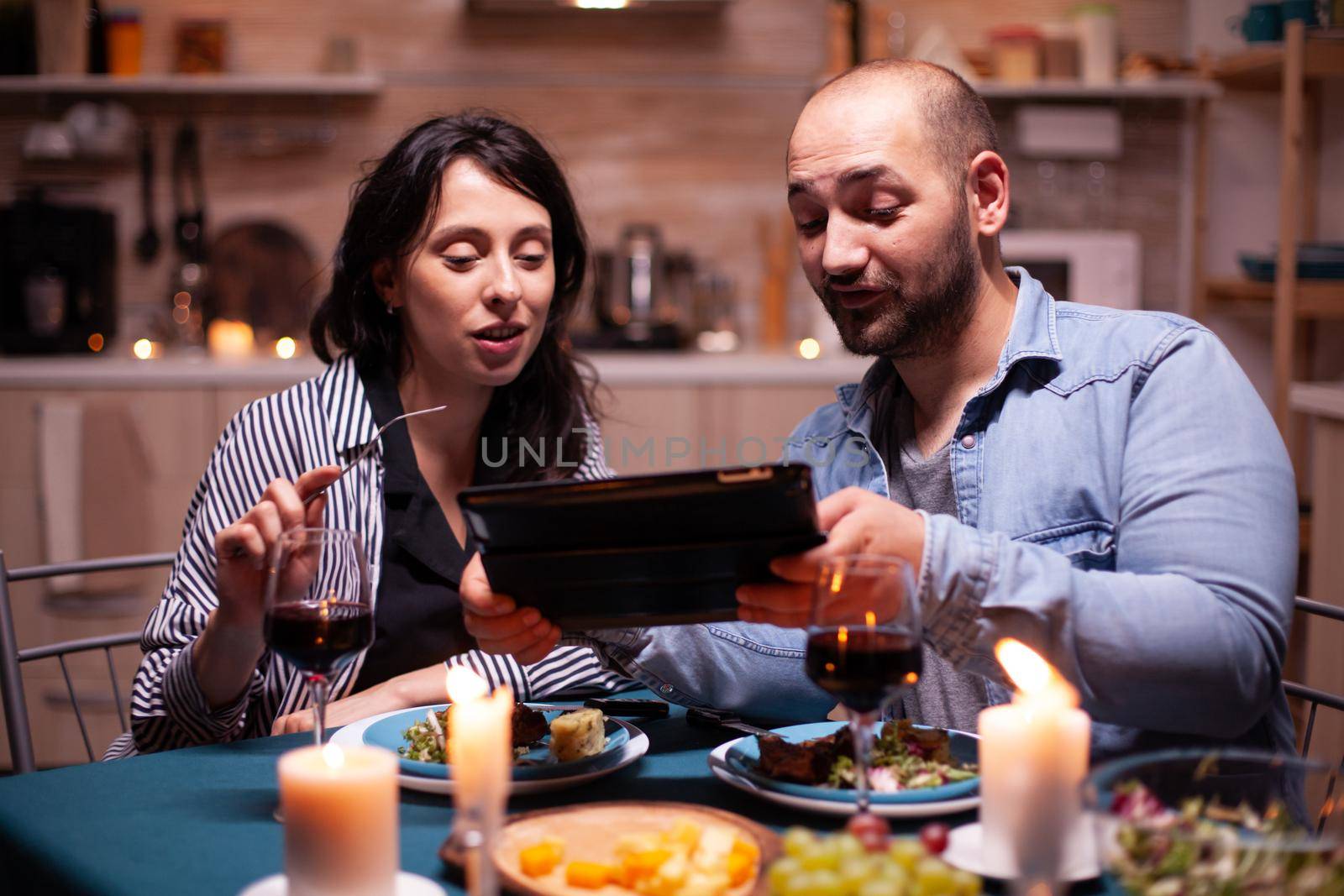 Cheerful young couple dining together using tablet during romantic dinner. Adults sitting at the table in the kitchen browsing, searching, , internet, celebrating, anniversary, happy., festive, enjoy.