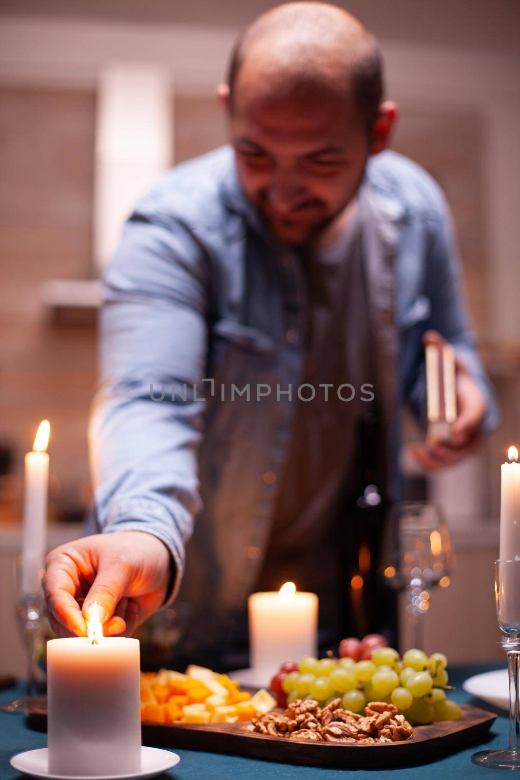 Lighting candle with matches. Husband preparing festive meal with healthy food for anniversary celebration, romantic date, sitting near the table in kitchen.