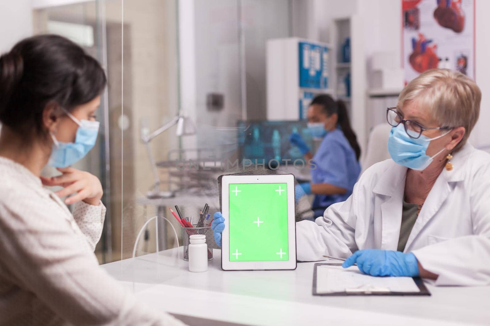 Doctor with face mask looking at tablet with green screen during consultation with sick patient. Nurse wearing blue uniform.