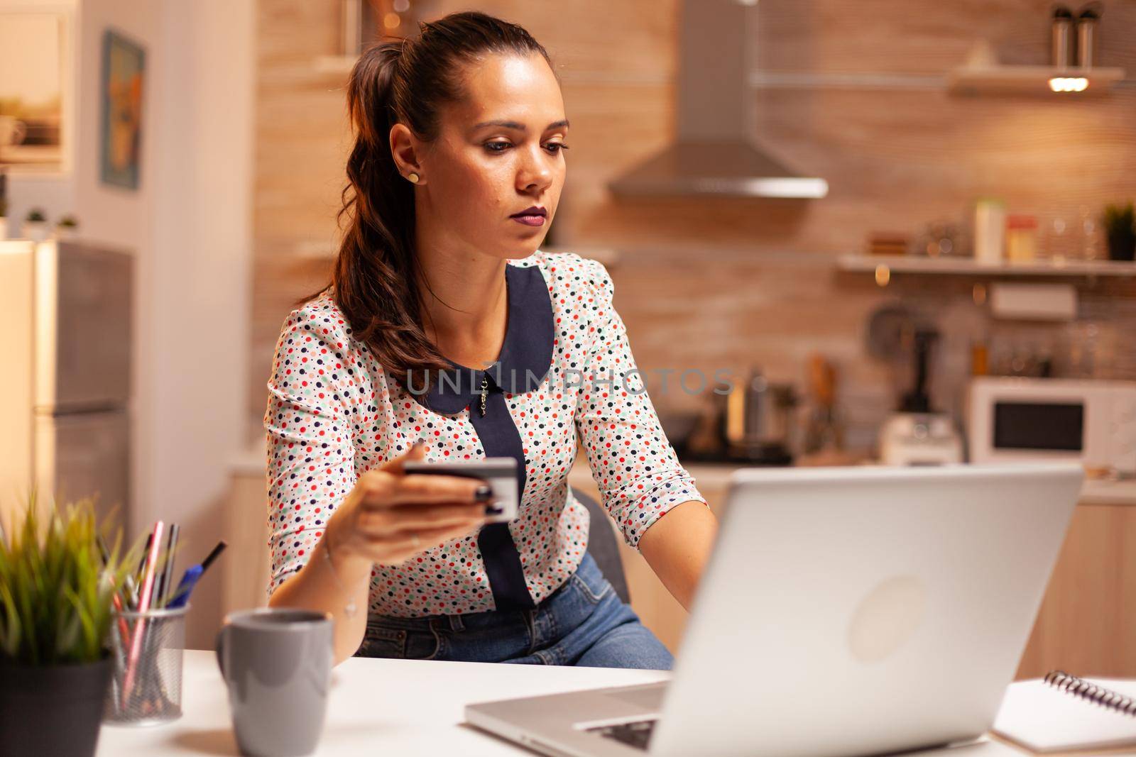 Woman making online payment in home kitchen late at night holding credit card. Creative lady doing online transaction using digital notebook conected to internet.