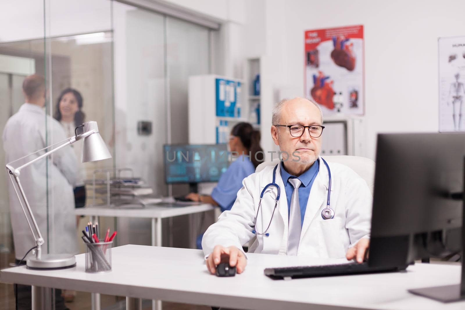 Senior specialist physician writing science report on computer in private clinic while his young colleague is looking at patient in hospital corridor wearing white coat.