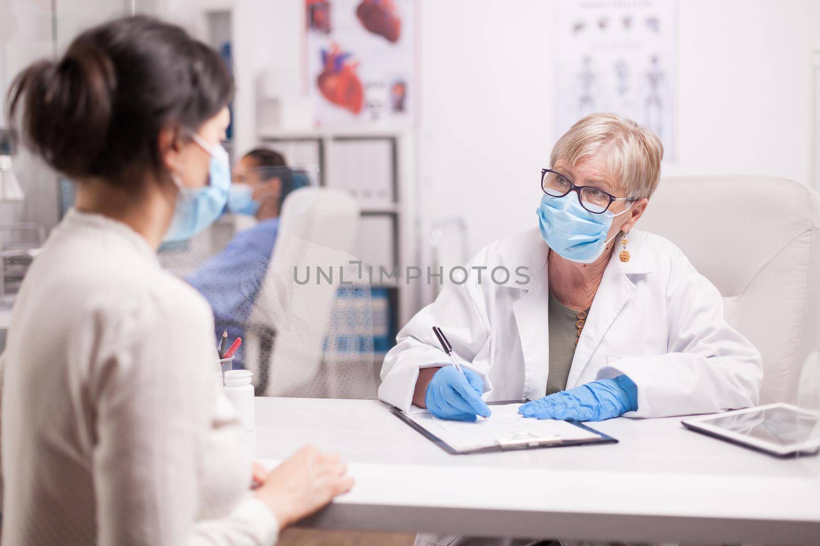 Doctor wearing protection mask against covid taking notes during consultation with patient in medical clinic. Nurse wearing blue uniform while working on computer.