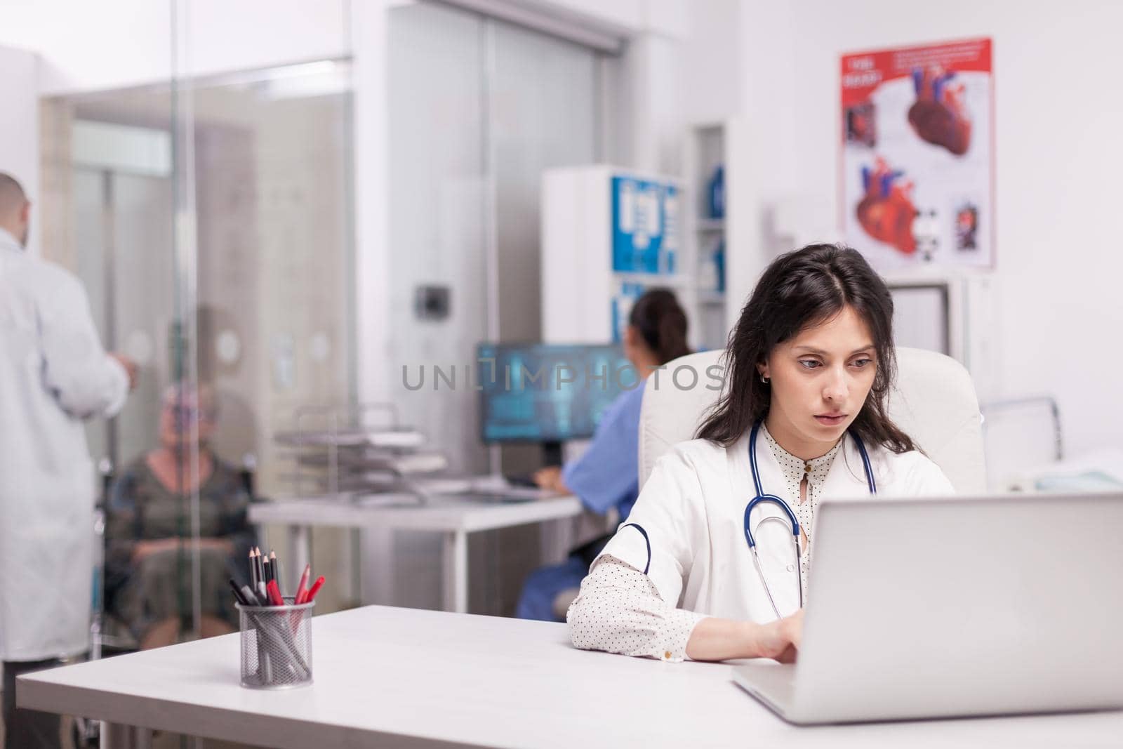 Young doctor in hospital office reading report about new drug to cure cancer on laptop. Disabled senior woman discussing with doctor wearing white coat on clinic corridor.