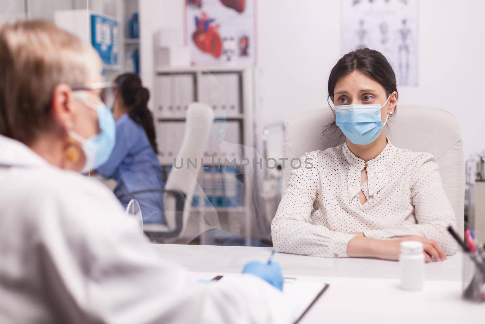 Stressed young patient with face mask against covid-19 during consultation with senior doctor in hospital office. Terrified woman.