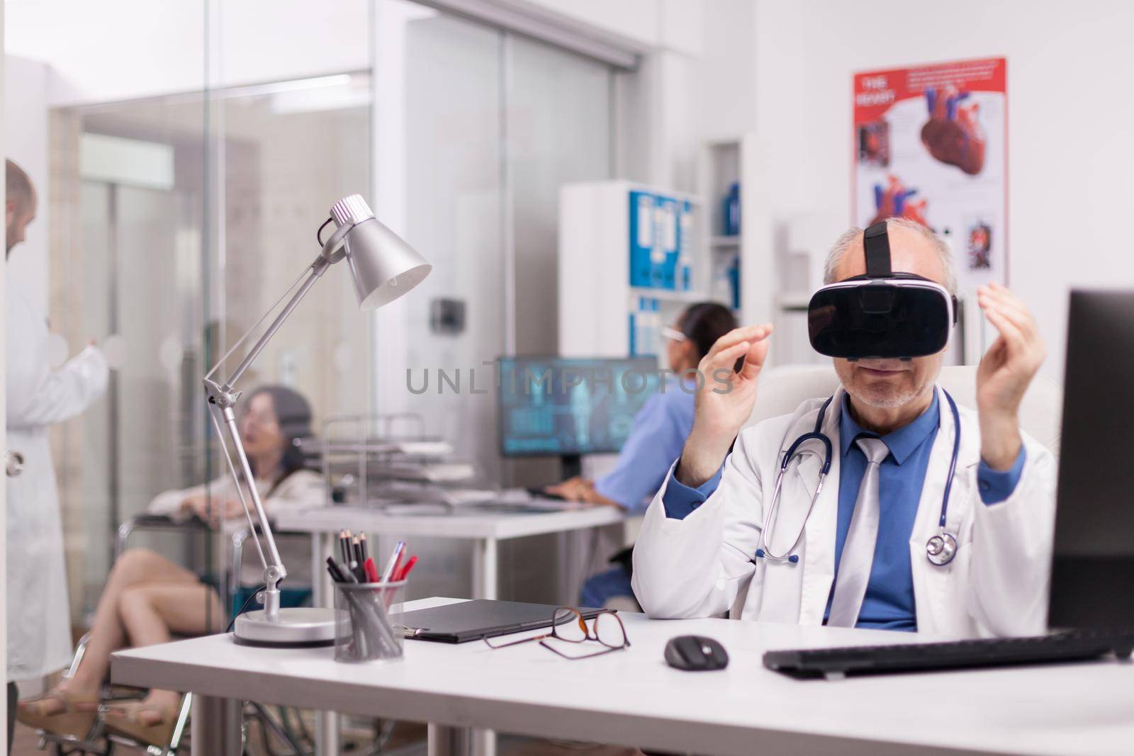 Senior doctor wearing virtual reality headset in hospital office while young therapist is discussing with disabled woman in wheelchair and nurse in blue uniform.