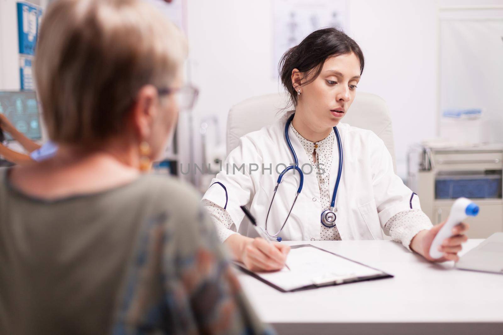 Medic writing senior patient diagnosis after measuring body temperature with thermometer gun in hospital office. Doctor with stethoscope wearing white coat.