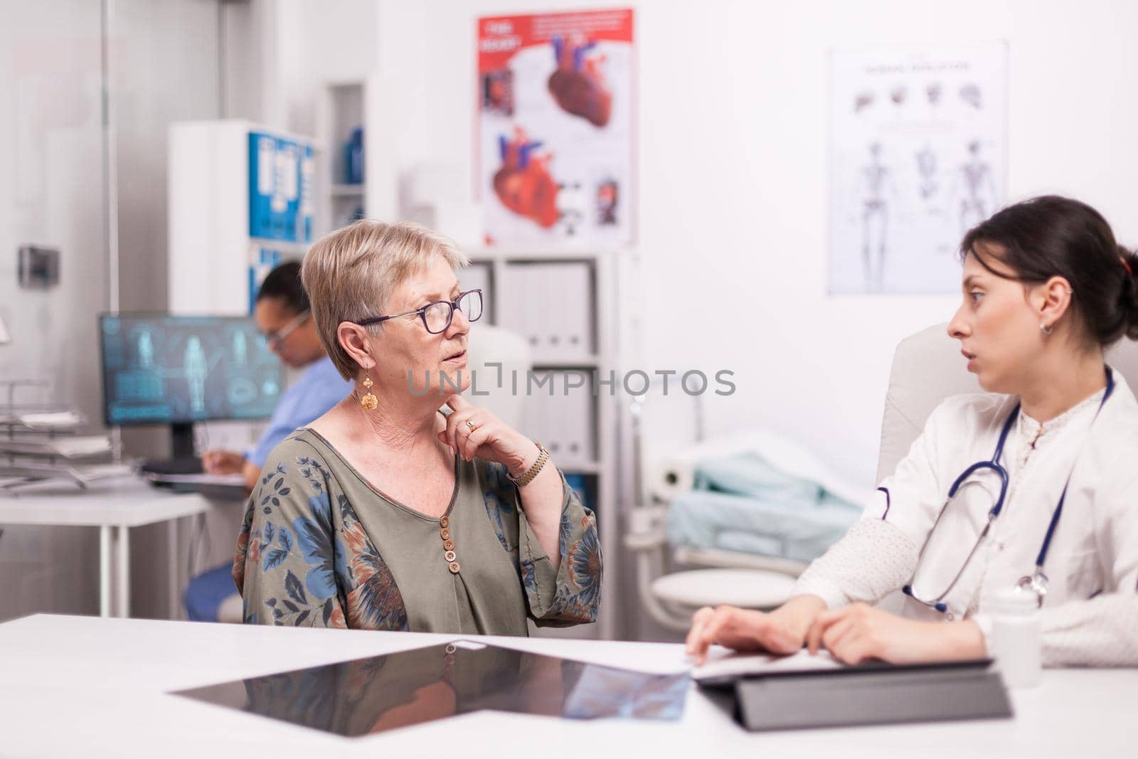Senior woman discussing about neck pain with doctor during consultation in hospital office. Medic wearing white coat and stethoscope. Mature woman with thyroid disorder.
