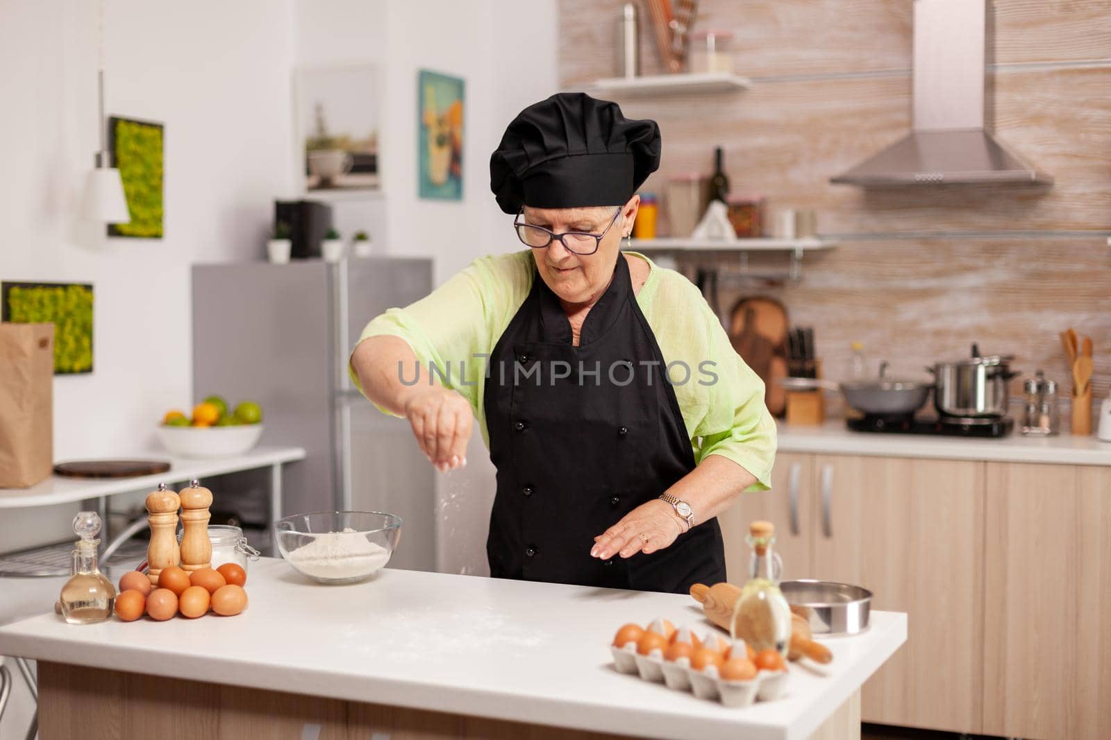 Elderly chef with uniform sprinkling flour in home kitchen wearing apron and bonette. Happy elderly chef with uniform sprinkling, sieving sifting raw ingredients by hand baking homemade pizza.