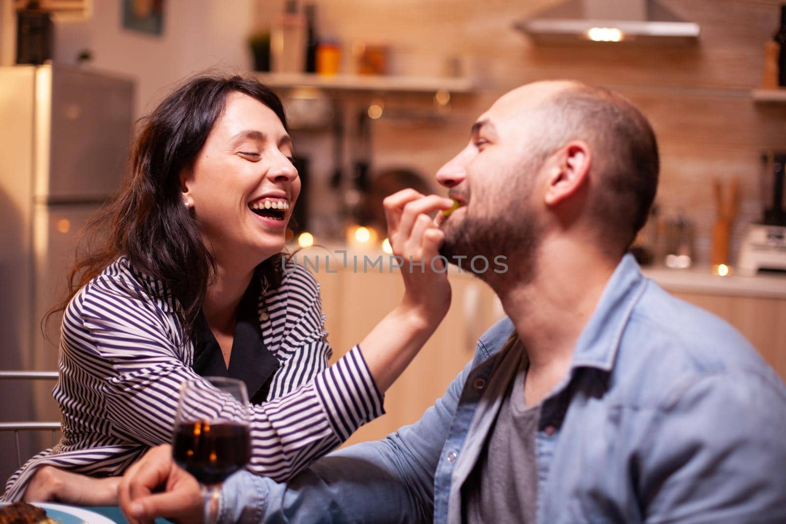 Husband feeding wife with grapes during romantic dinner celebrating relationship. Wife and husband celebrating anniversary with red wine, tender moments at candle lights in kitchen.