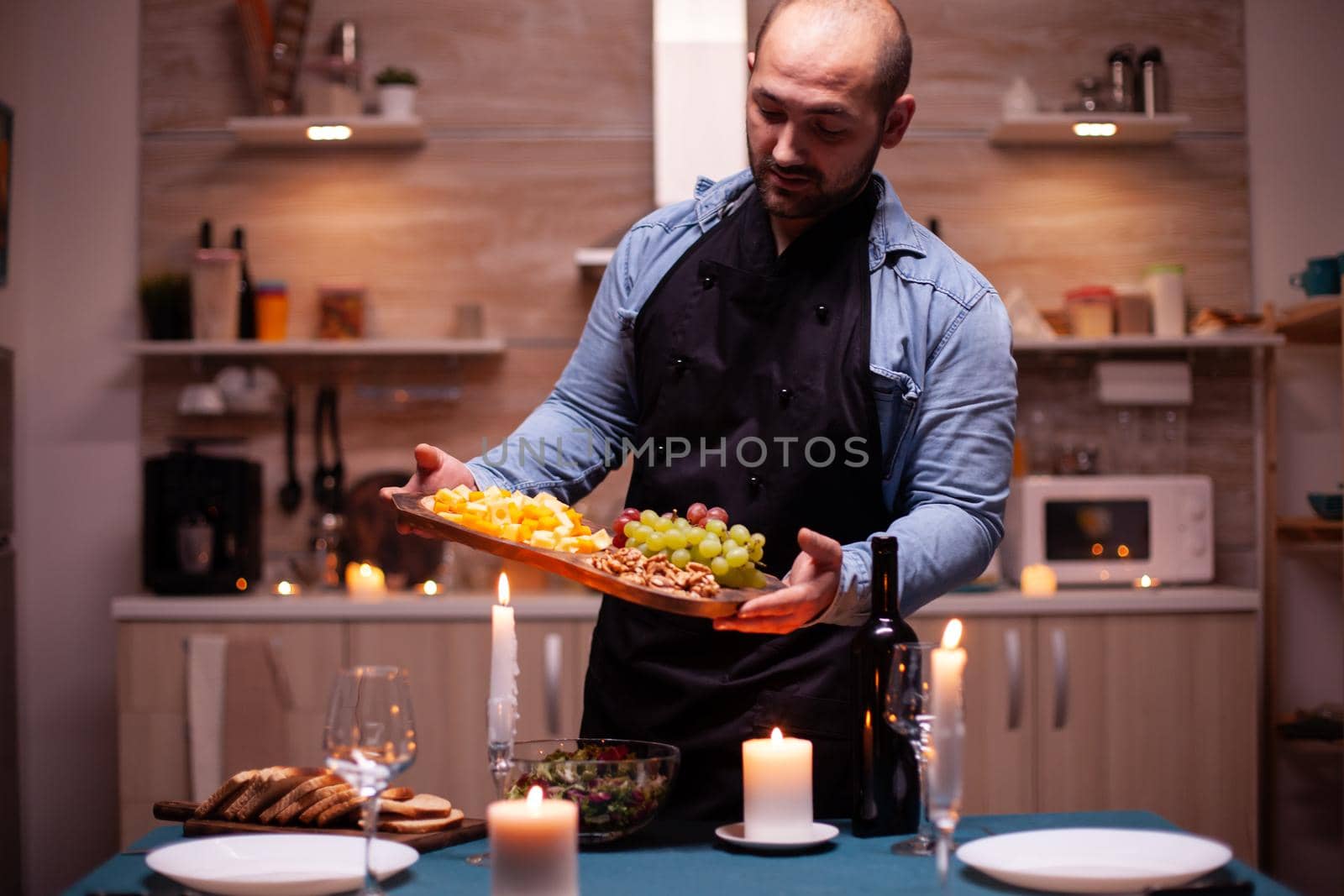 Man wearing apron holding wooden plate with grapes and cheese in kitchen waiting wife for dinner. Man preparing festive dinner with healthy food, cooking for his woman a romantic dinner,