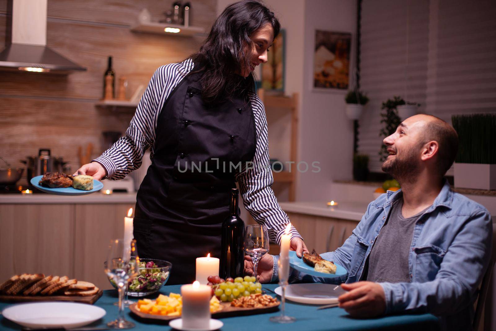 Couple celebrating relationship with romantic dinner in dining room. Woman preparing festive dinner with healthy food, cooking for his man romantic dinner, talking, sitting at table