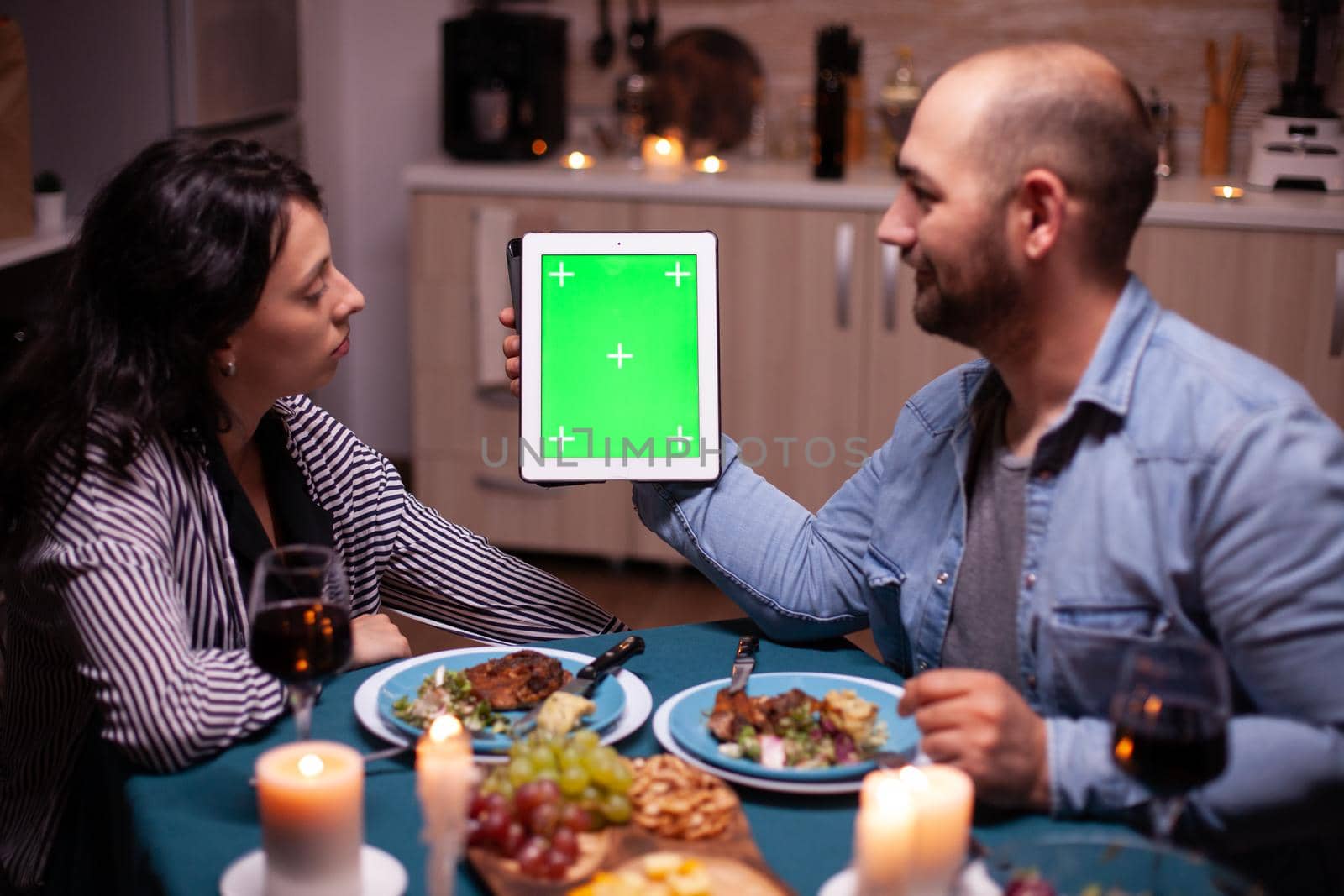 Husband holding tablet with green screen and looking at wife during romantic dinner.