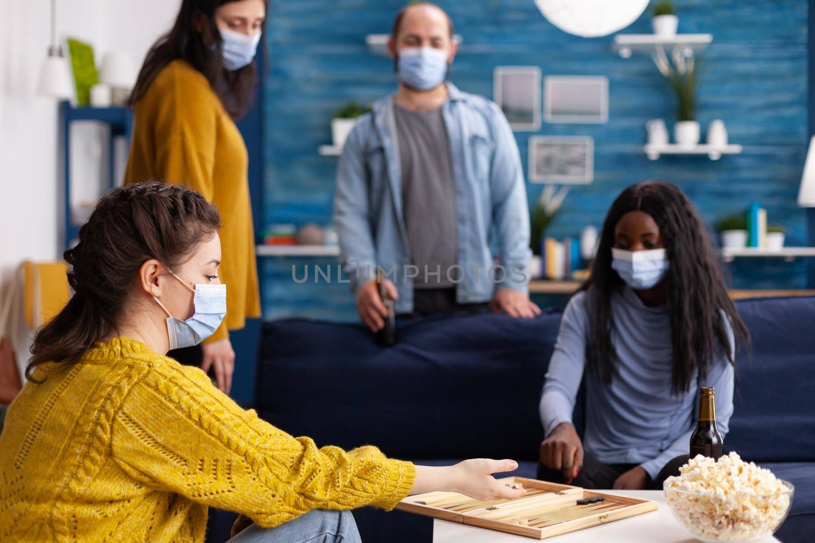 Cheerful happy woman using dice playing backgammon with multiethnic friends wearing face mask as prevention for covid 19 spread during global pandemic sitting on couch drinking beer and eating popcorn.