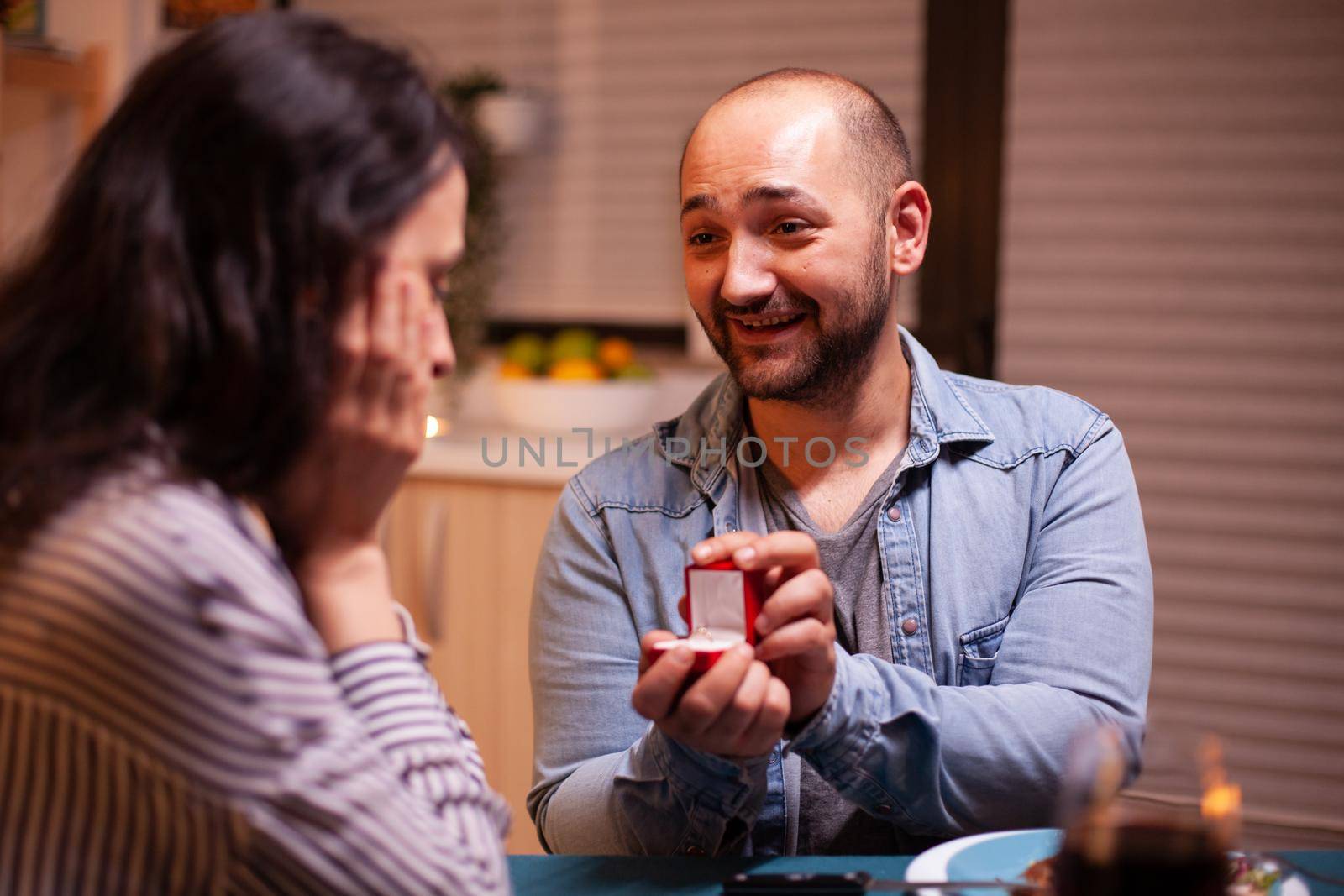 Young man asking wife to marry him during dinner in kitchen. Man making proposal to his girlfriend in the kitchen during romantic dinner. Happy caucasian woman smiling being speechless