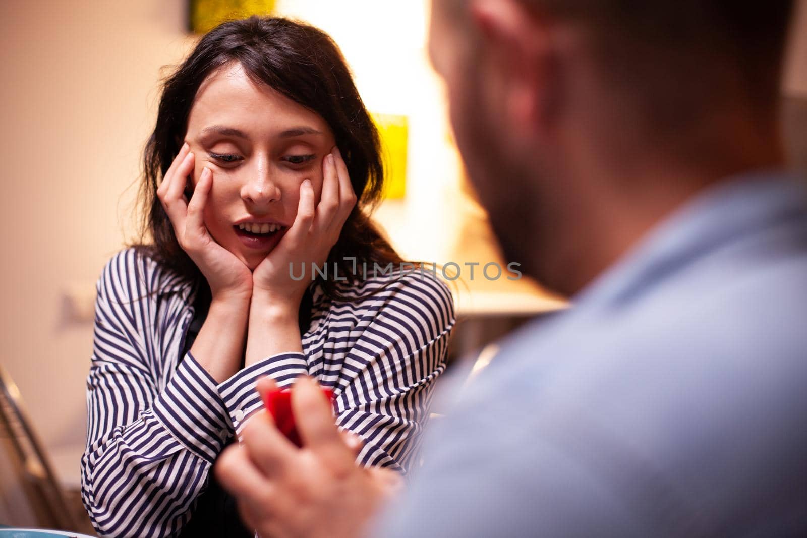 Woman looking happy at engagement ring during marriage proposal while having romantic dinner. Man asking his girlfriend to marry in the kitchen during romantic dinner. Happy caucasian woman smiling being speechless