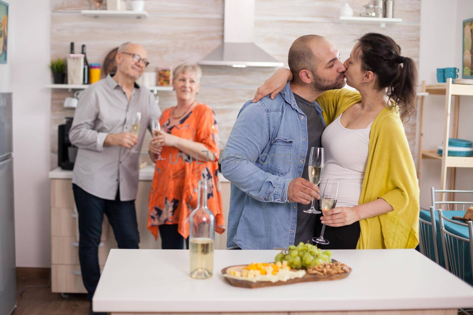 Young man kissing wife in from of his parents during brunch in kitchen. Assorted cheese and grapes on wooden plateu.