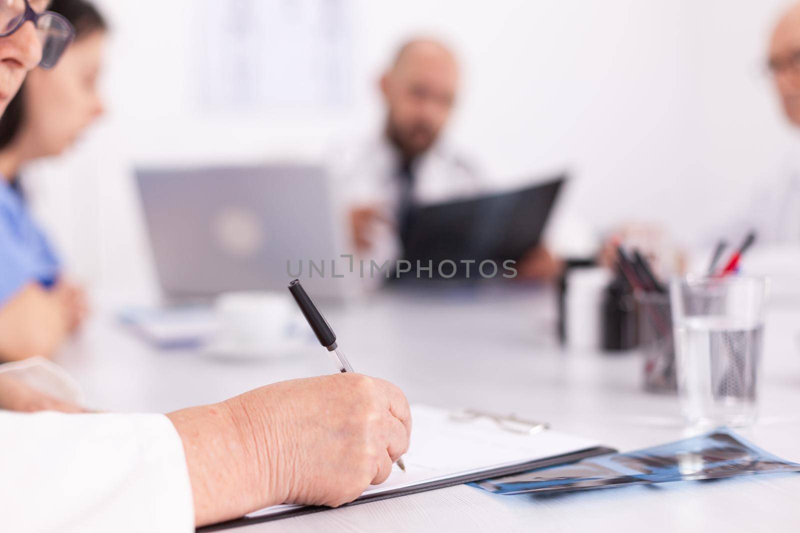 Close up of unknown doctor taking notes on clipboard during medical briefing in conference room. Clinic expert therapist talking with colleagues about disease, medicine professional