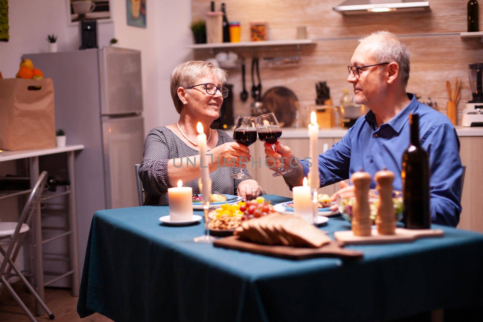 Candles burning on kitchen table and old couple toasting with glasses for red wine for their relationship. Happy cheerful senior elderly couple dining together in the cozy kitchen, enjoying the meal.