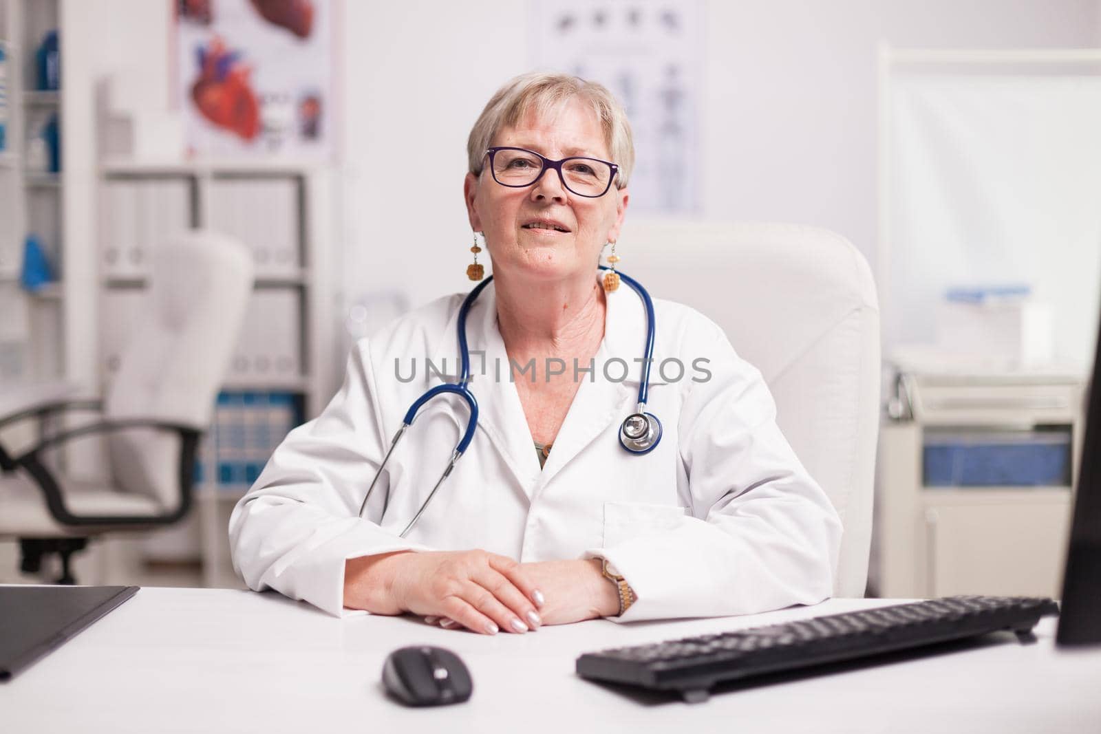 Senior medical practitioner with stethoscope in hospital cabinet smiling to camera.