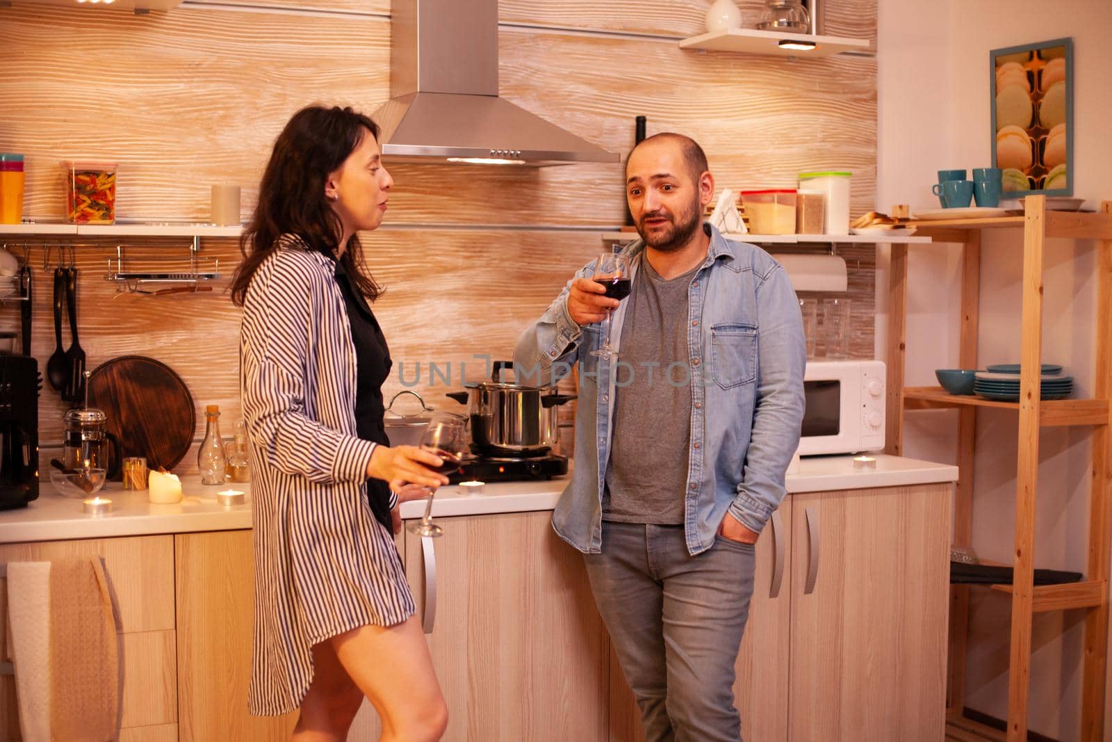 Lovers drinking wine and talking in kitchen during the evening. Adult couple at home, having a conversation, smiling, enjoying the meal in dining room.