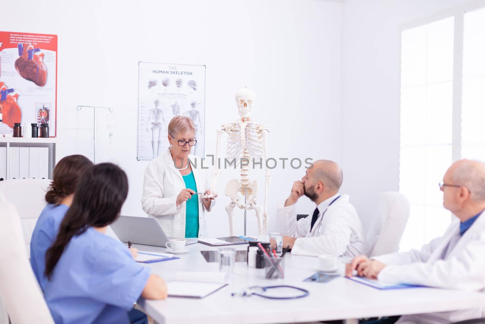 Mature surgeon doing demonstration on human anatomy on skeleton for medical staff in conference room. Clinic expert therapist talking with colleagues about disease, medicine professional