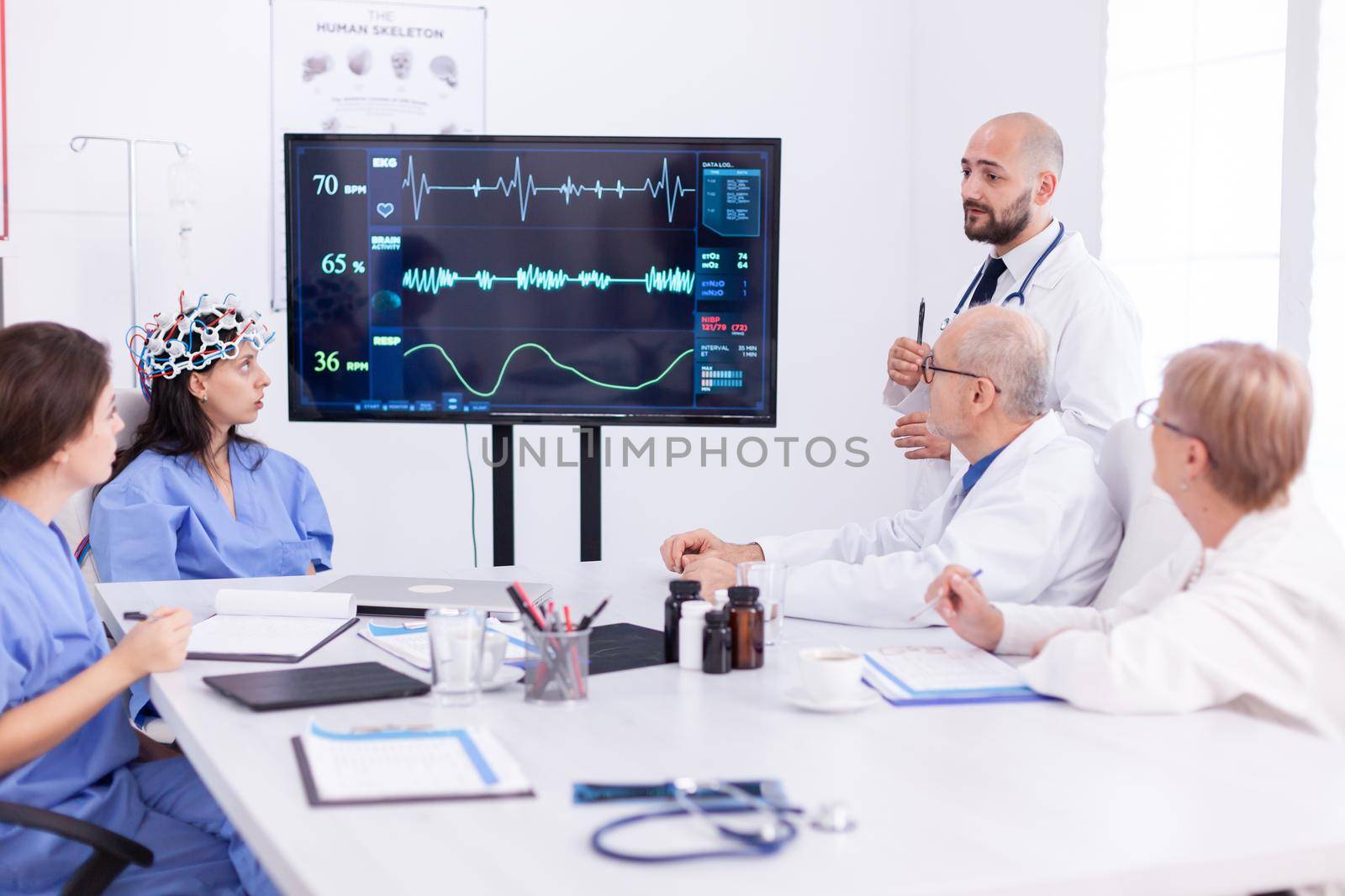 Nurse wearing scanner for brain waves during medical experiment for neuoroscience conference. Monitor shows modern brain study while team of scientist adjusts the device.