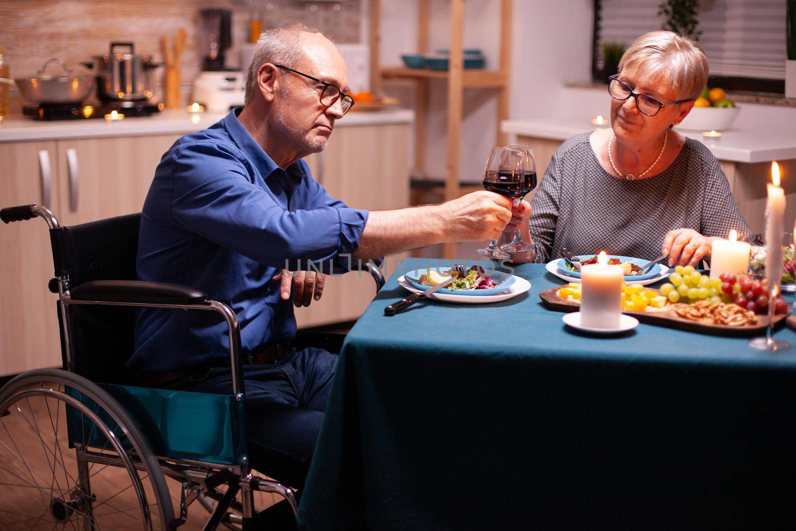Man with disabilities having dinner with wife and clinking wine glass. Wheelchair immobilized paralyzed handicapped man dining with wife at home, enjoying the meal