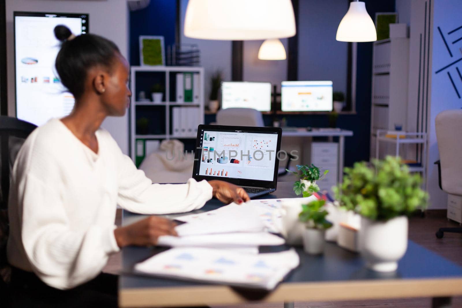 African woman working late at night looking at charst on laptop. Focused employee doing overtime for job respecting deadline of project writing, searching.