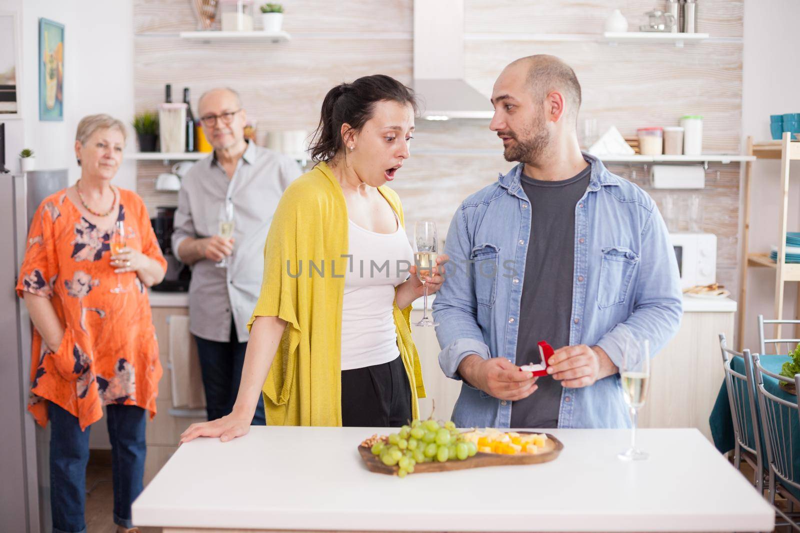 Man proposing wife during family lunch. Shocked young woman looking at engagement ring while holding a glass of wine