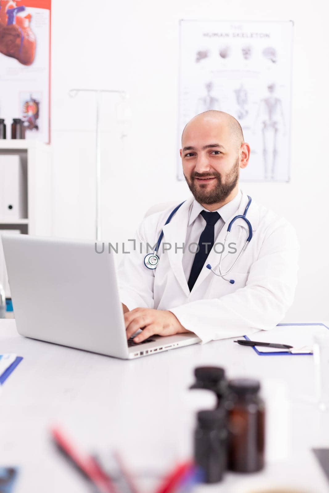Portrait of doctor smiling holding hands on laptop keyboard looking at camera in hospital office wearing lab coat. Medical practitioner using notebook in clinic workplace, expertise, medicine.