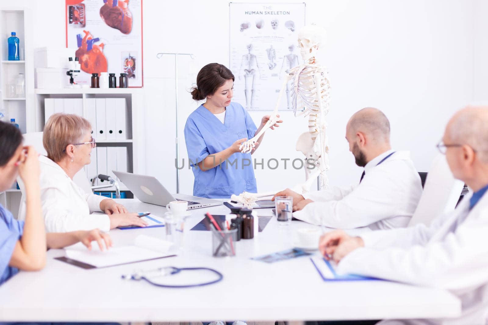 Female nurse demonstrating on skeleton in front of medical surgeons in conference room. Clinic expert therapist talking with colleagues about disease, medicine professional