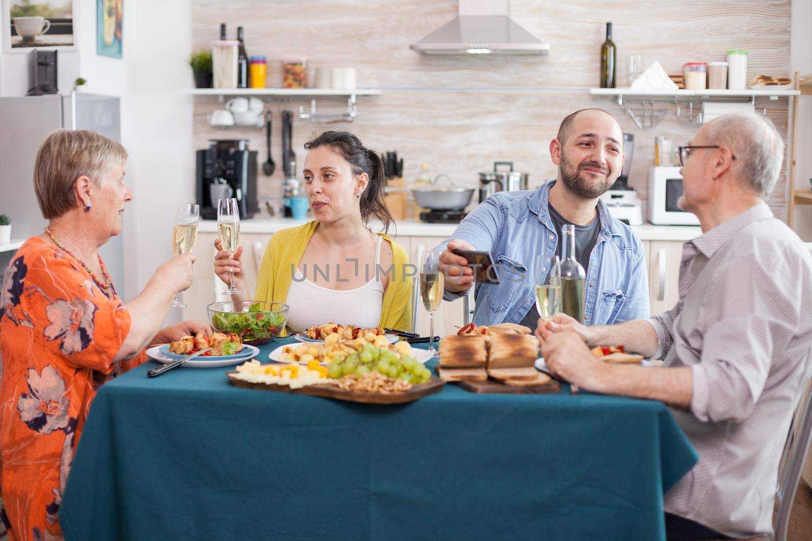 Son and senior father looking at smartphone during family lunch. Mother and daughter holding wine glasses. Delicious seasoned potatoes.