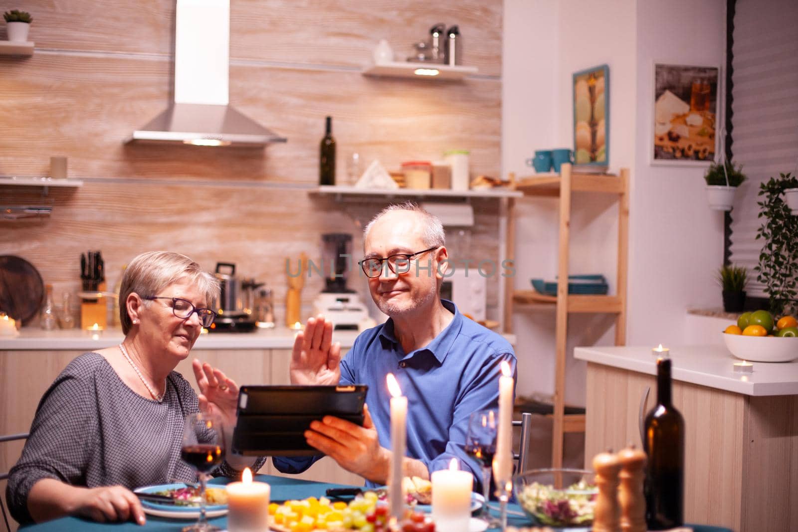 Mature couple saying hello during video conference in kitchen while having romantic dinner using tablet pc. Couple sitting at the table, browsing, talking, using internet,in dining room.