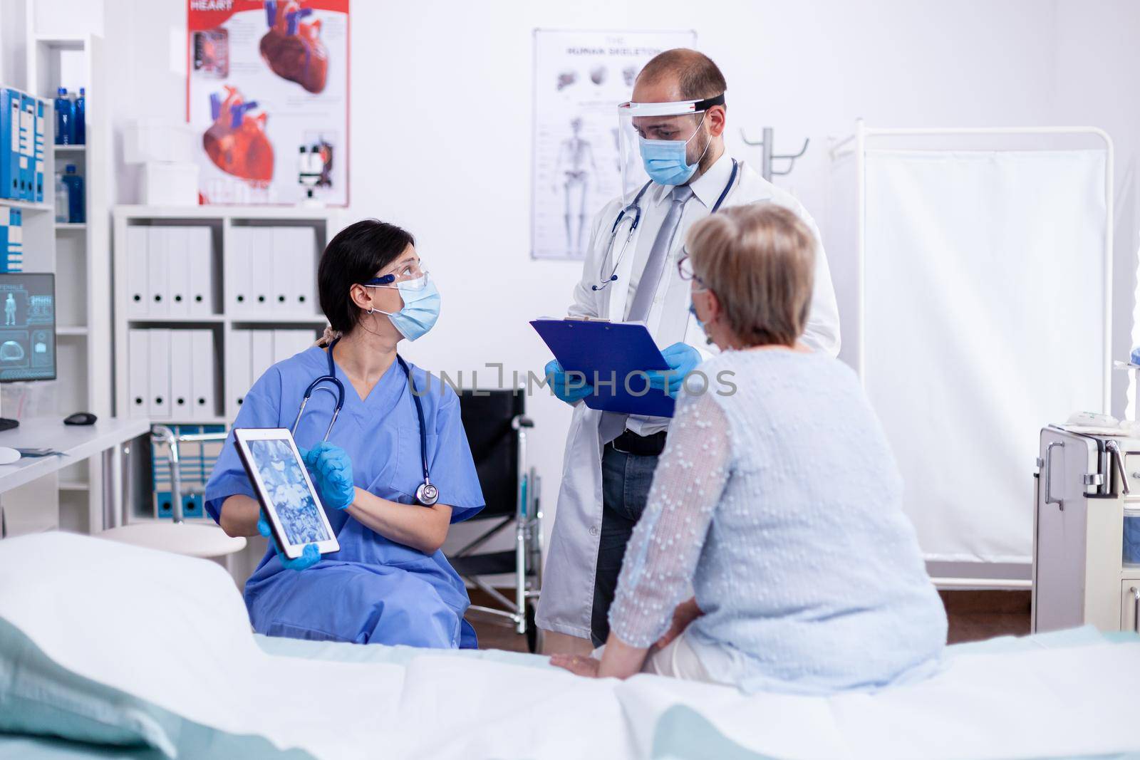 Nurse consulting with doctor during examination of senior woman in hospital room holding tablet pc and wearing face mask against covid-19 as safety precaution. Medical control for infections,