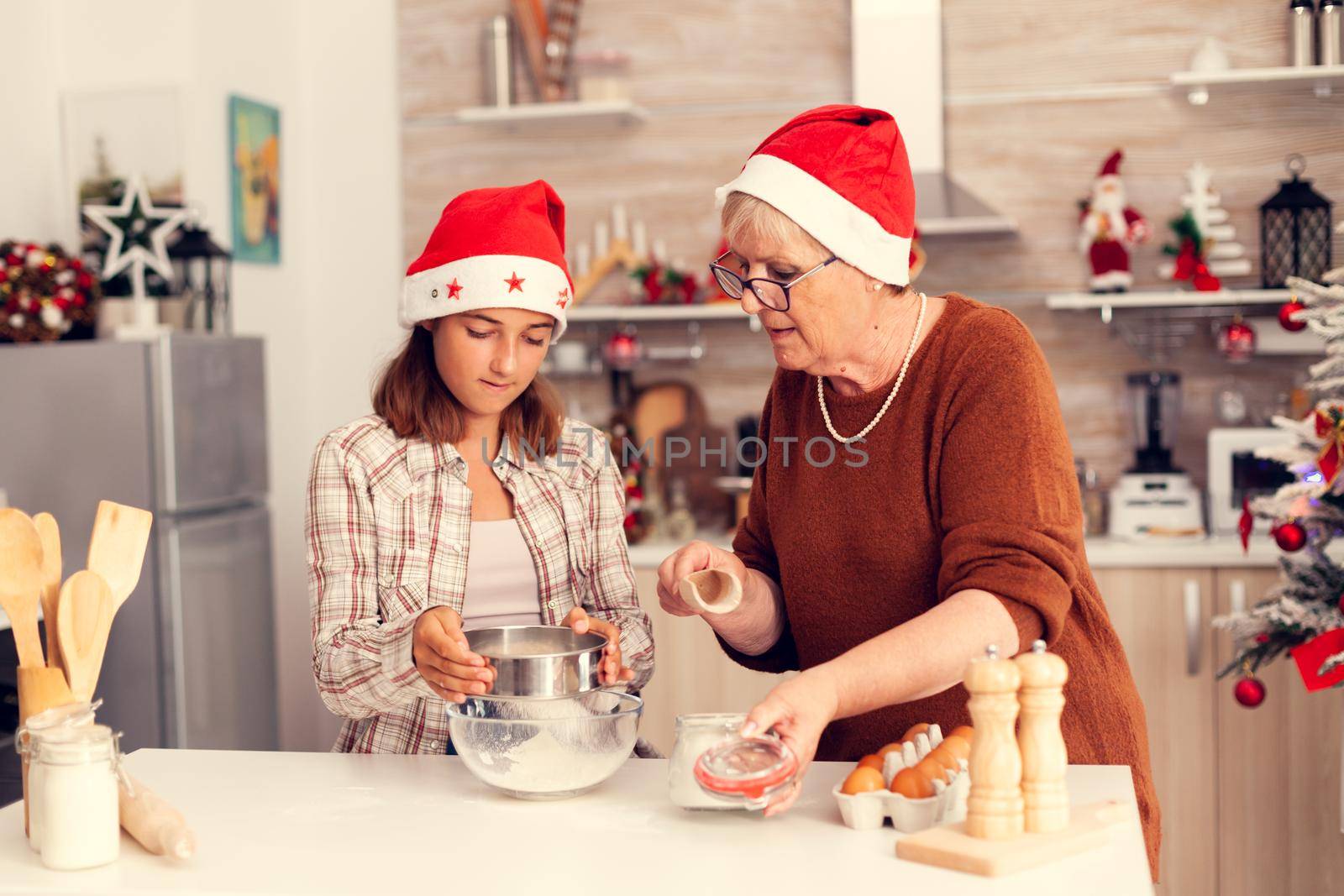 Granddaughter and mother on christmas day doing cookies with love. Happy cheerful joyfull teenage girl helping senior woman preparing sweet biscuits to celebrate winter holidays wearing santa hat.