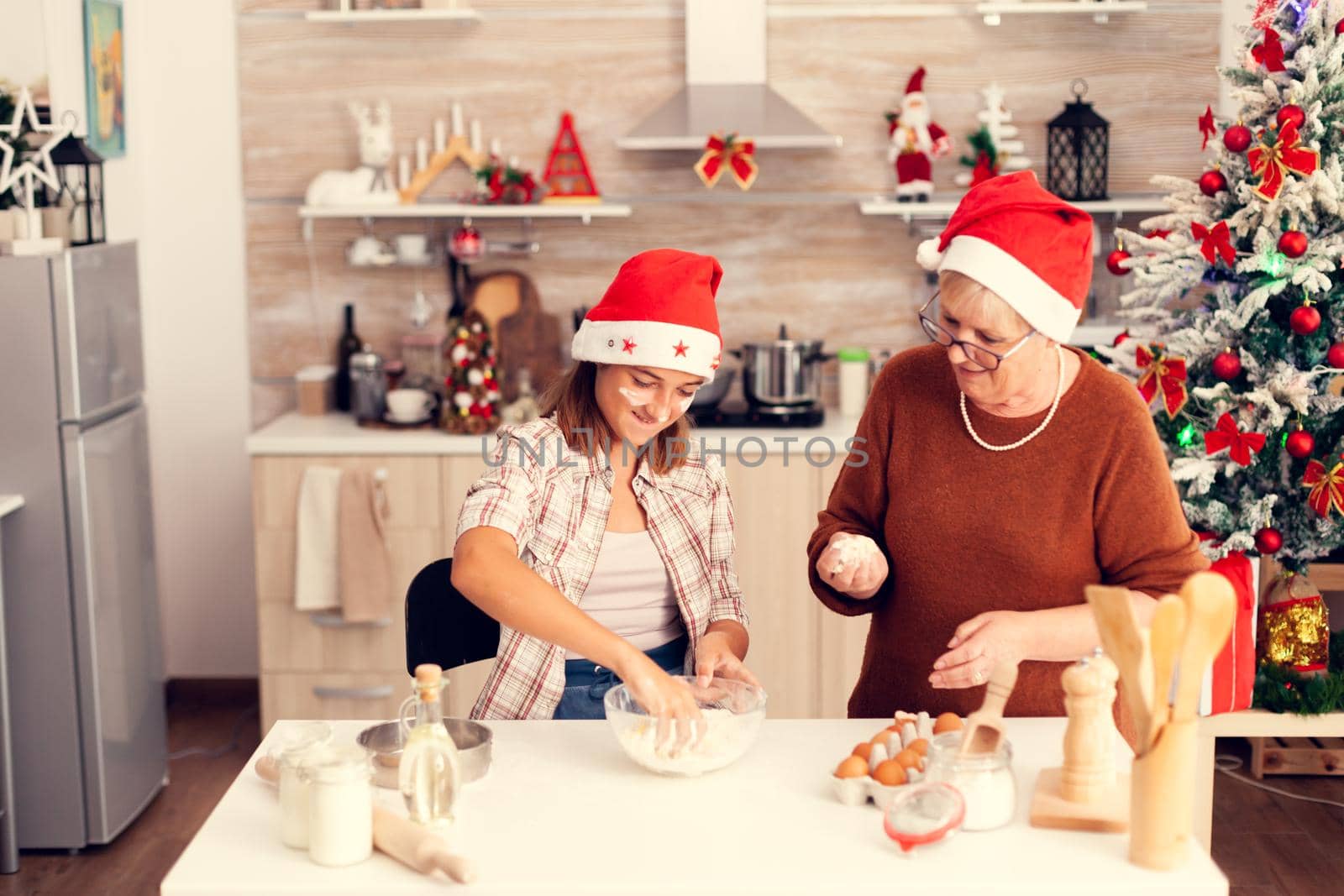 Joyfull kid mixing dough on christmas day. Happy cheerful joyfull teenage girl helping senior woman preparing sweet cookies to celebrate winter holidays wearing santa hat.