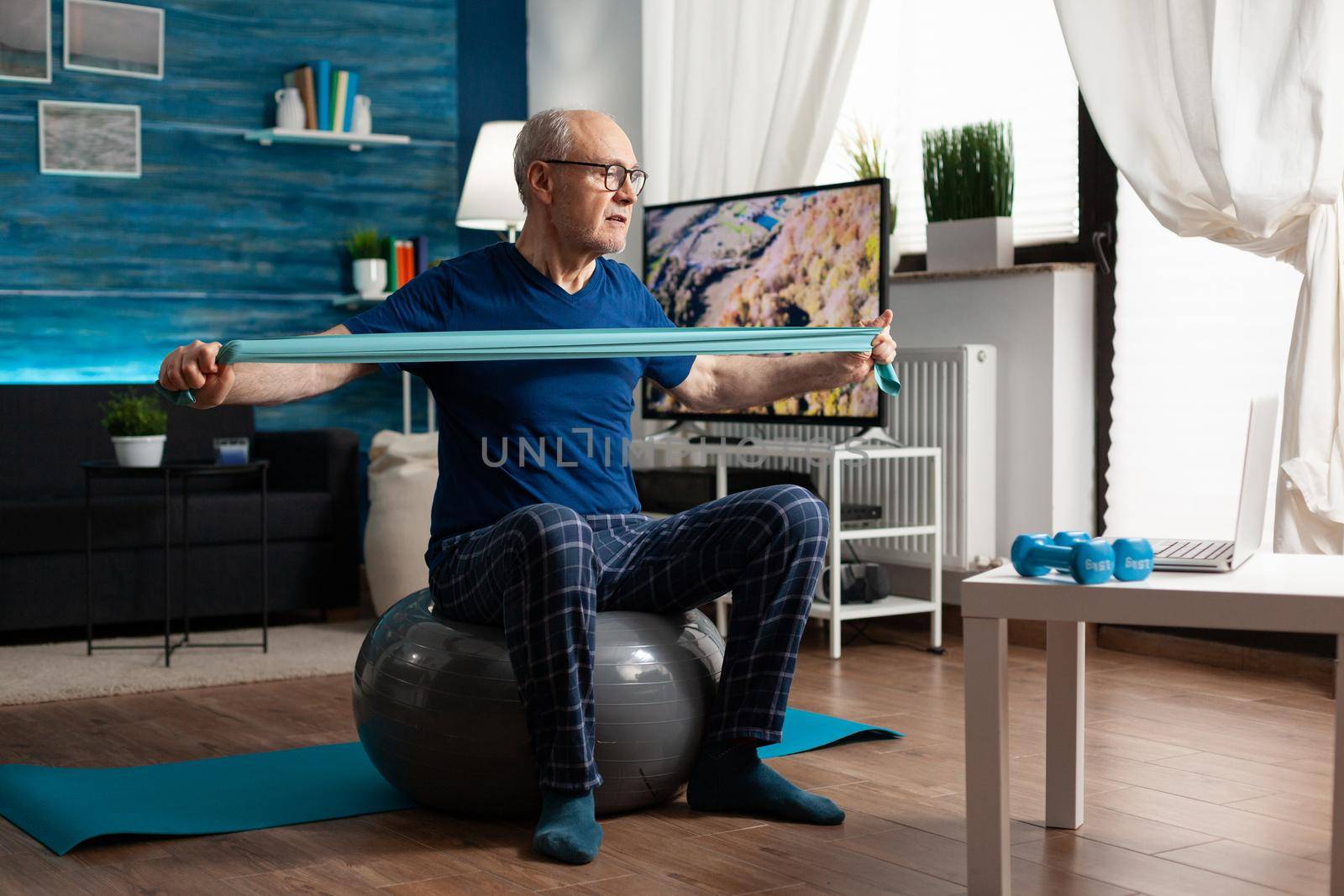 Invalid pensioner practicing active arm exercises using resistance elastic band watching online fitness workout video on laptop. Pensioner working body healthcare sitting on swiss ball in living room