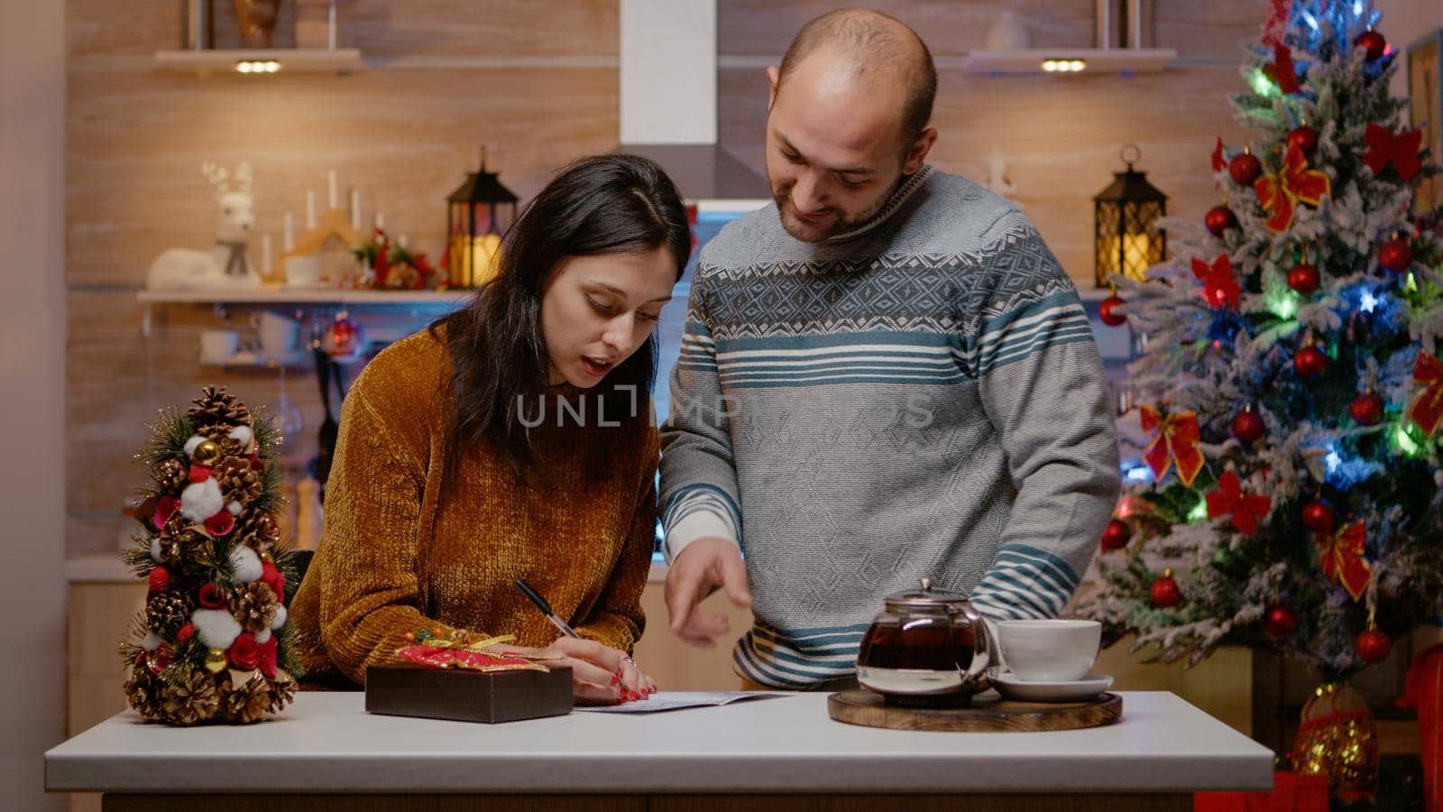 Cheerful couple preparing gift and christmas card to give present box to relatives for holiday festivity. Festive man and woman signing postcard on christmas eve for seasonal celebration.