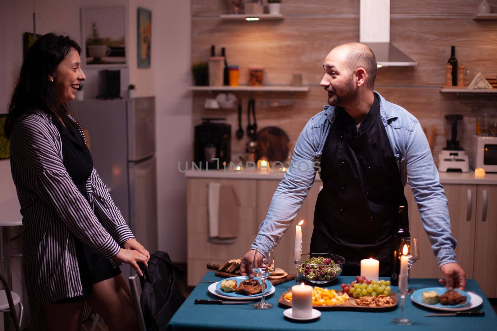 Cheerful husband surprising wife with dinner in dining room after she arrives from work. Man preparing festive dinner with healthy food, cooking for his woman a romantic dinner,