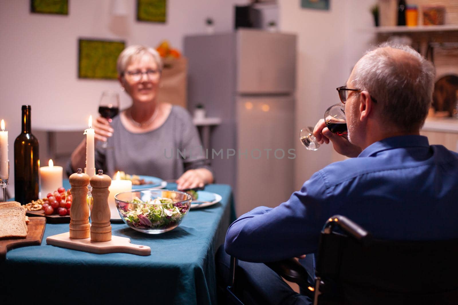 Paralyzed man in wheelchair drinking wine during dinner with wife. Happy cheerful senior elderly couple dining together in the cozy kitchen, enjoying the meal, celebrating their anniversary.