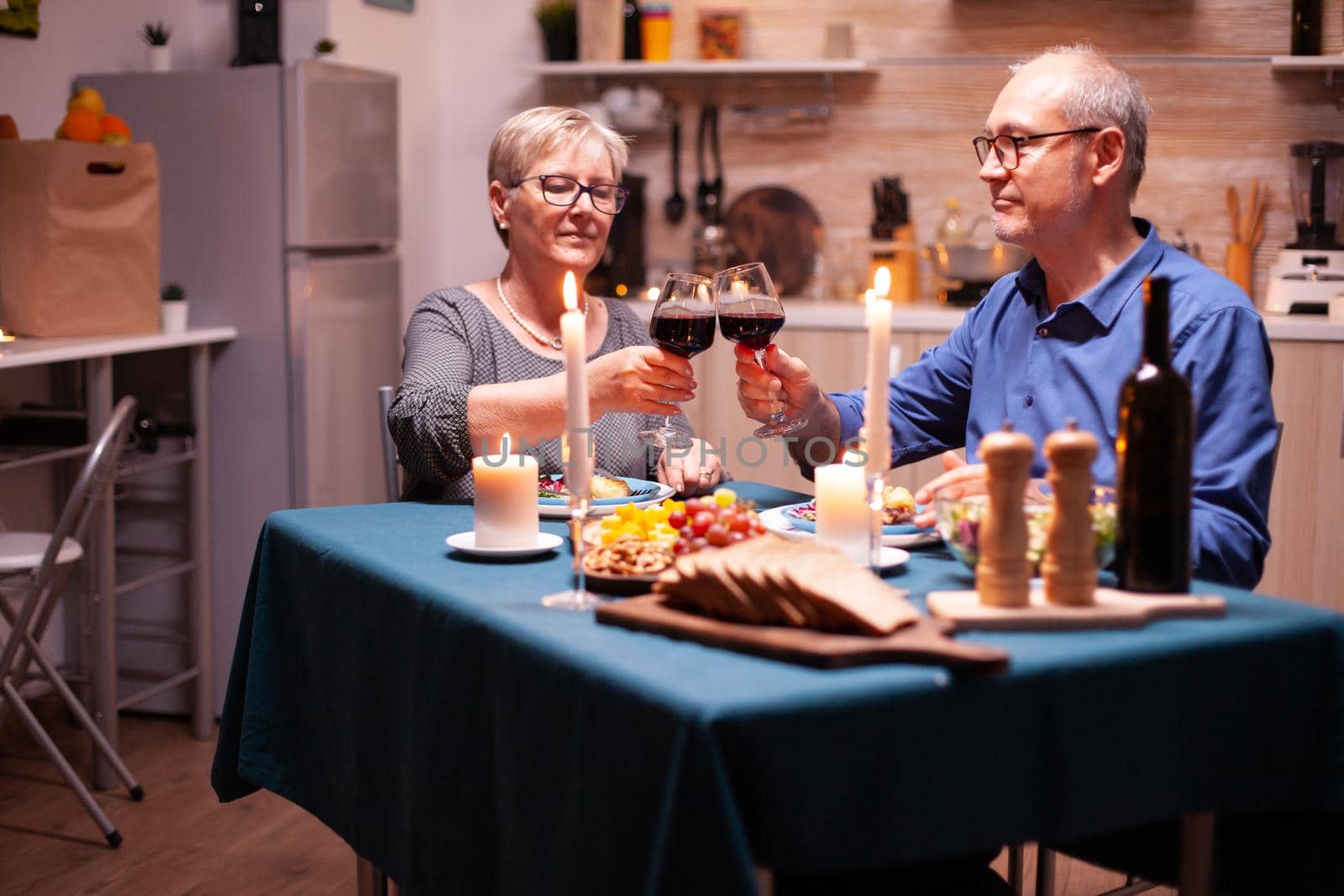 Elderly couple dining together with wine in kitchen. Happy cheerful senior elderly couple dining together in the cozy kitchen, enjoying the meal, celebrating their anniversary.