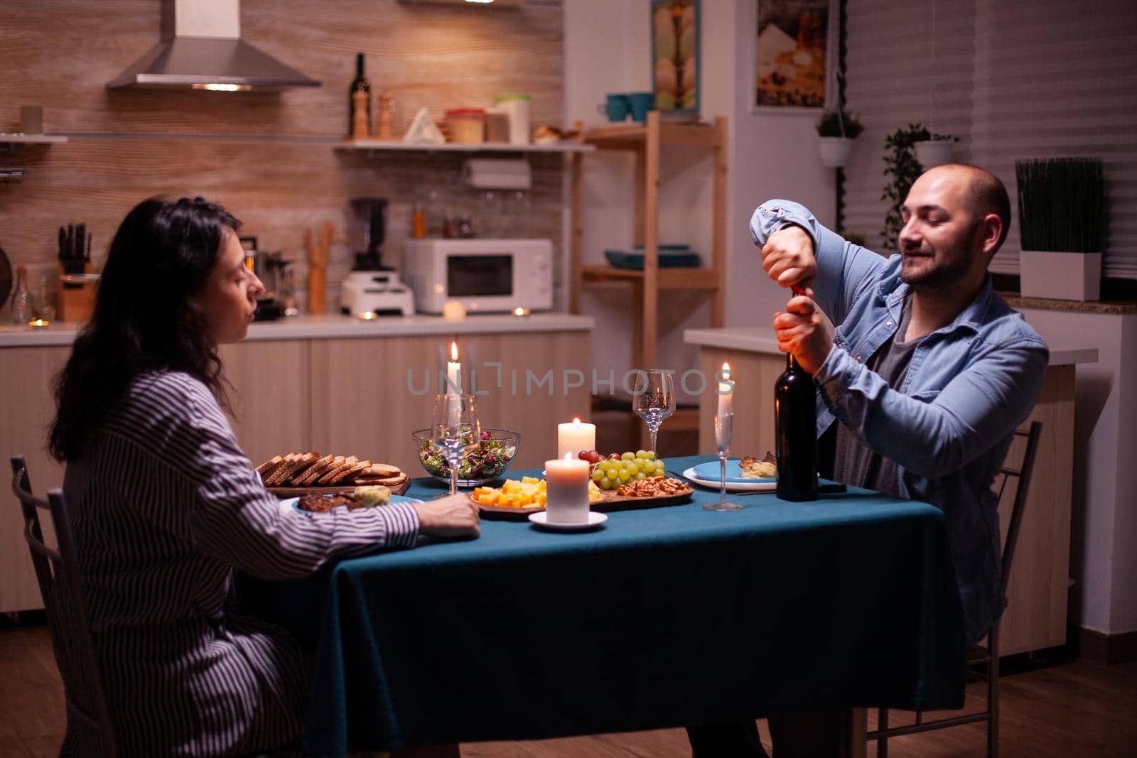 Boyfriend opening wine bottle. Happy couple talking, sitting at table in kitchen, enjoying the meal, celebrating their anniversary at home with healthy food